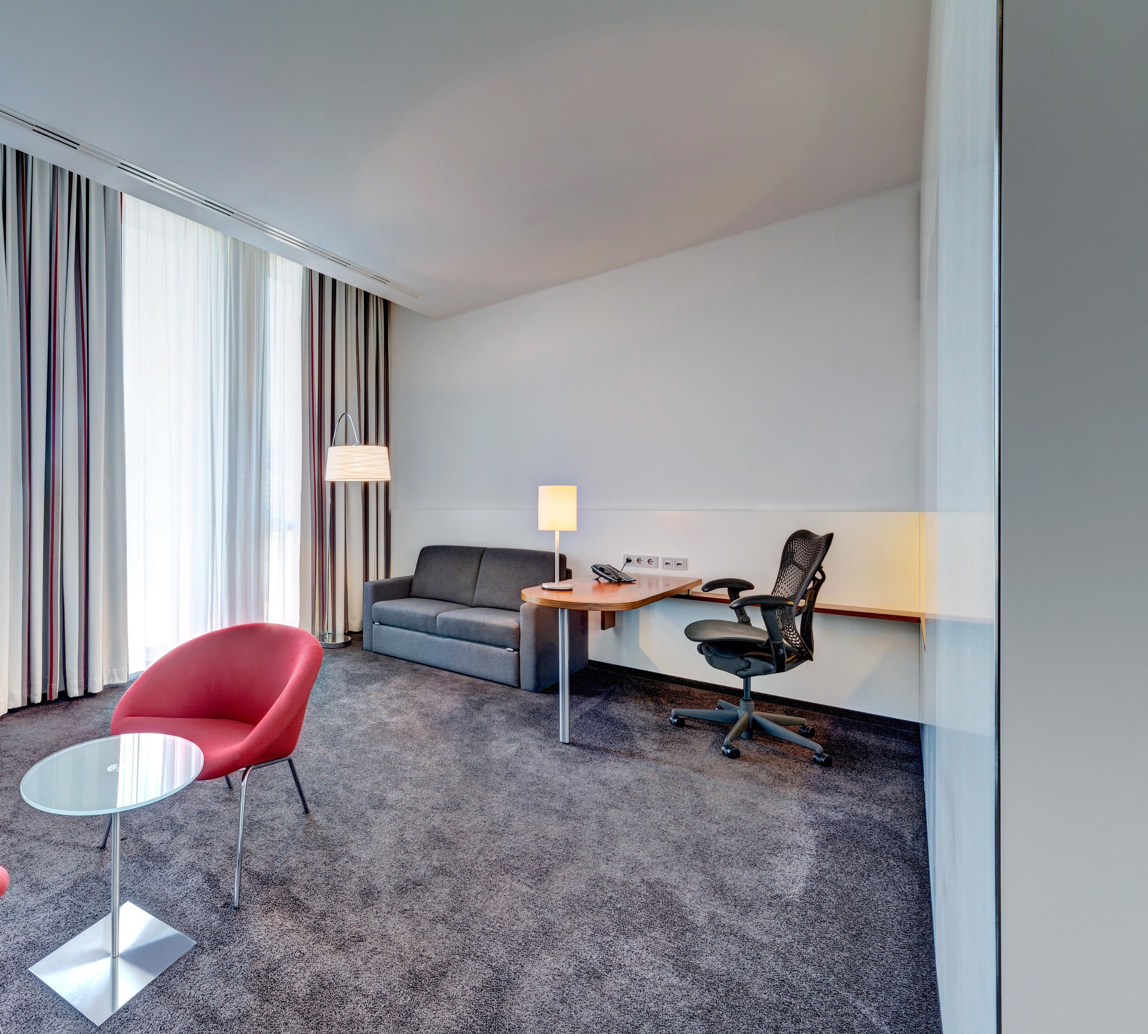 Une chambre d’hôtel avec un canapé gris, un fauteuil rouge, une petite table ronde, un fauteuil de bureau noir, une lampe et une fenêtre avec des rideaux.