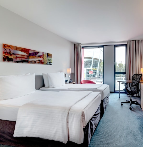 Habitación de hotel con dos camas, un escritorio, una silla y un ventanal con vistas a un edificio.