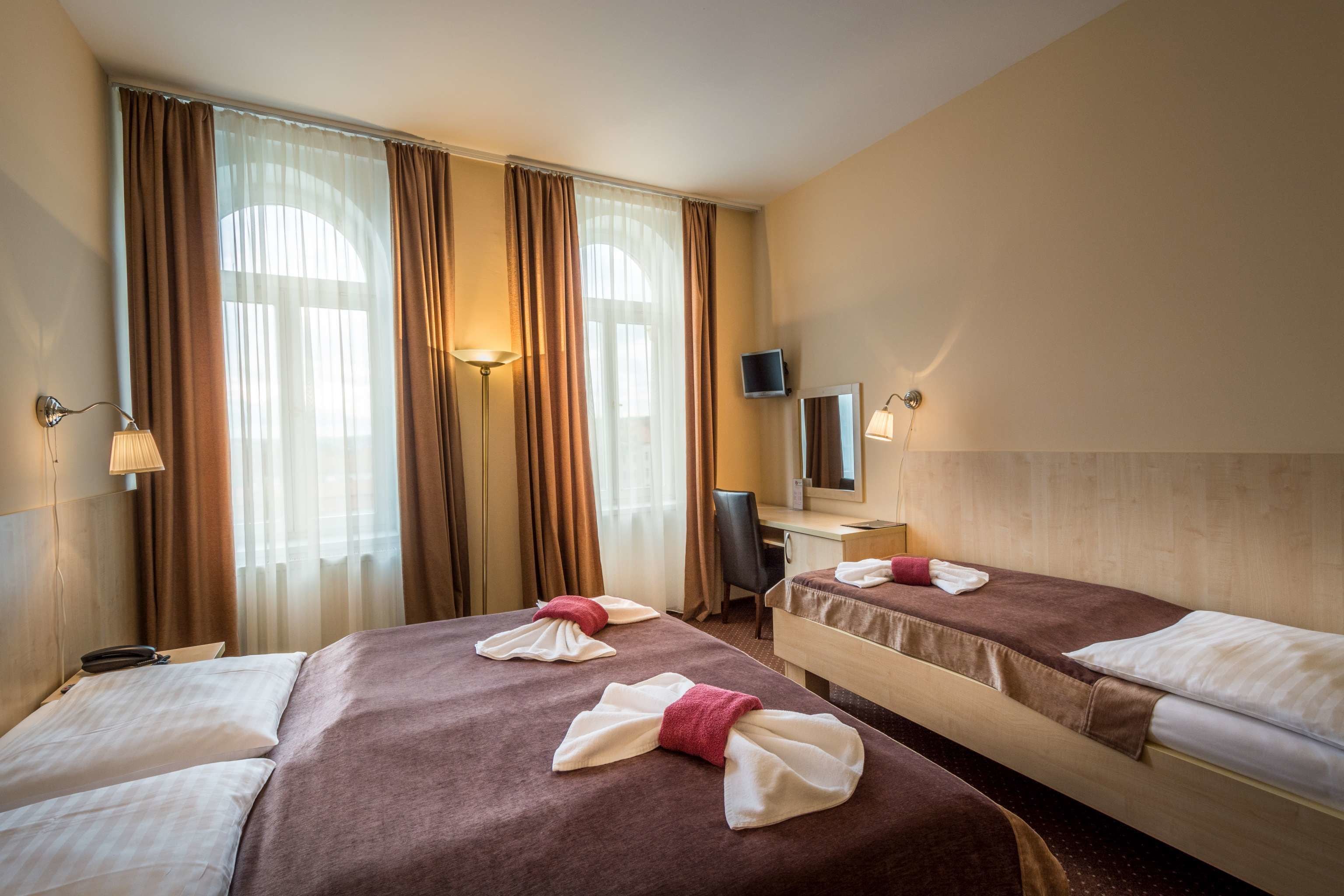 A hotel room with two beds, a desk, a chair, a TV, and a large window with curtains.