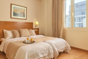 Quarto de hotel com uma cama grande, mesa de cabeceira, quadro na parede, janela com cortinas e bandeja com copo de suco de laranja.