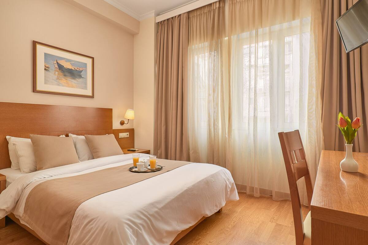 Una habitación de hotel con una cama grande, una mesita de noche con lámpara, un cuadro enmarcado en la pared y una ventana con cortinas.