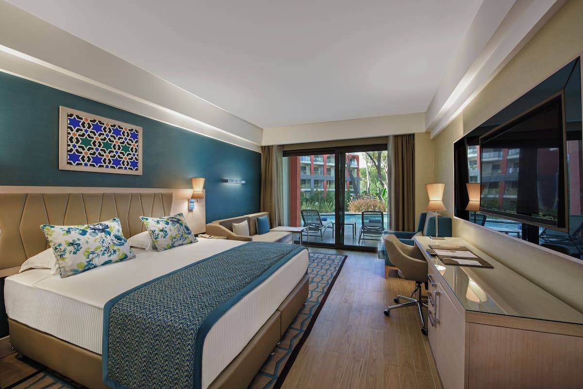 A hotel room with a large bed, a desk, a TV, and a balcony with outdoor seating.