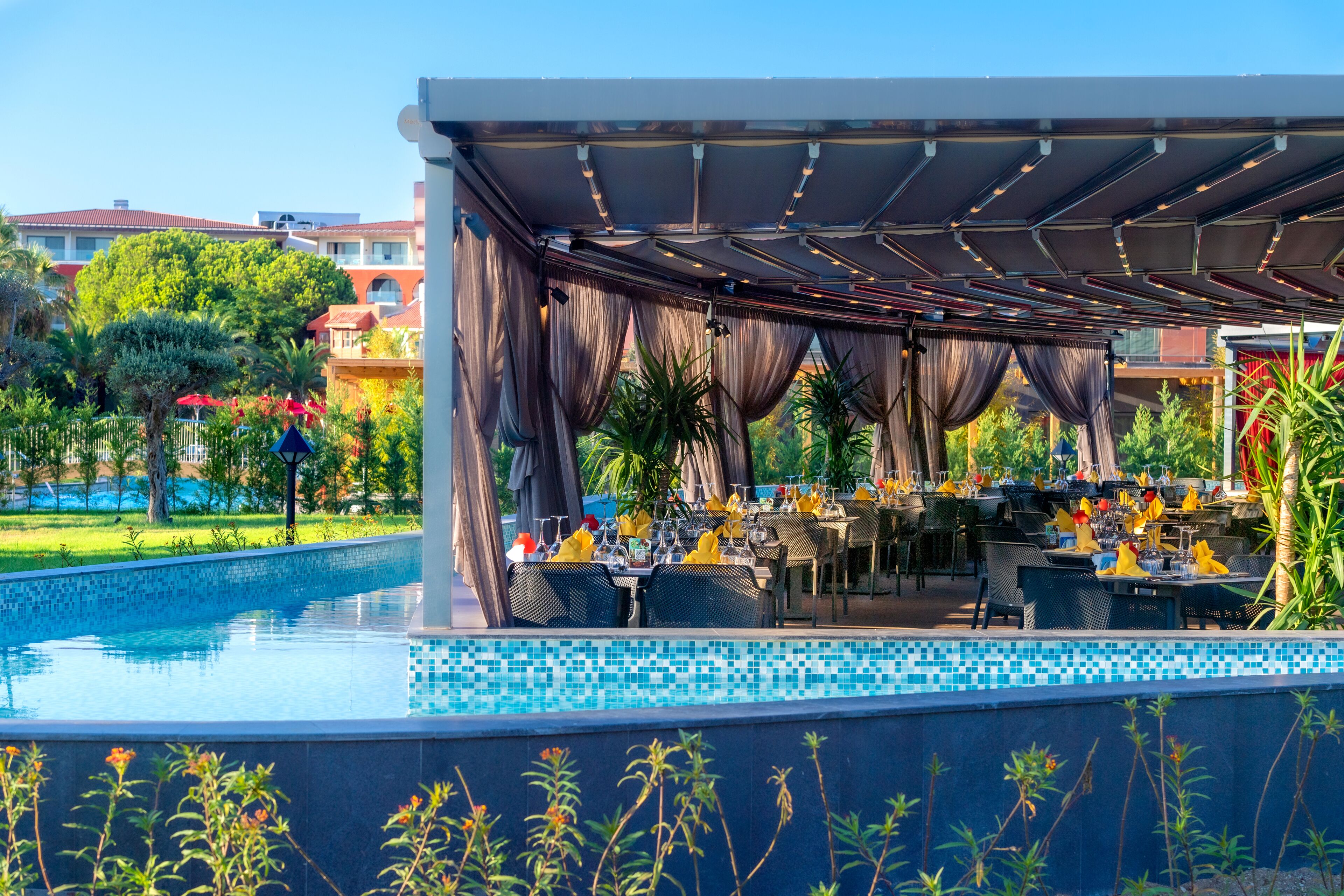 A poolside dining area with tables and chairs, a covered seating area, and a pool with a tiled edge.