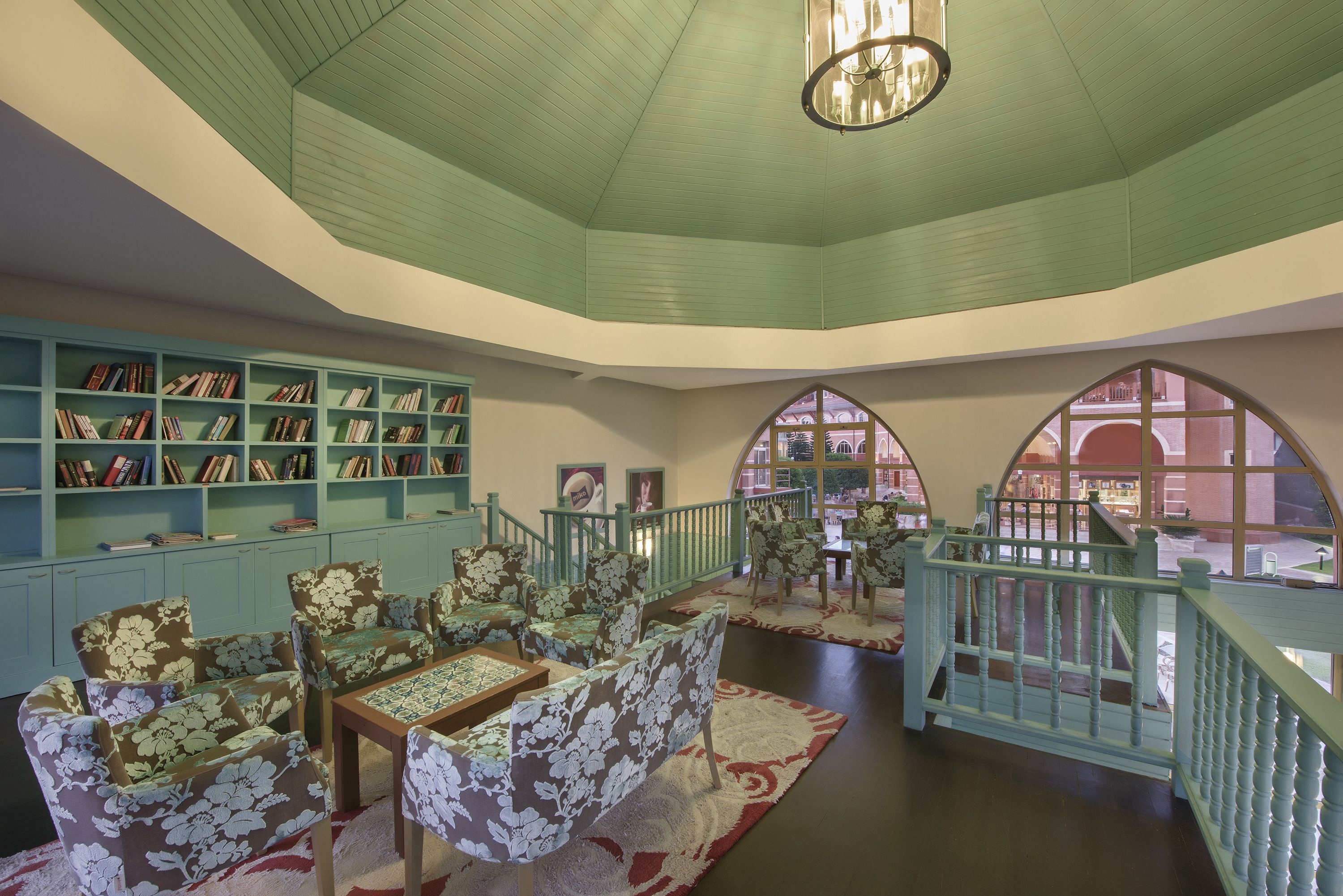 A library with bookshelves, floral-patterned chairs, a table, and a staircase with a view outside.