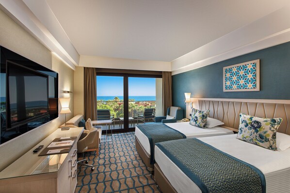 A hotel room with a large TV, two beds, a desk with a chair, and a view of the ocean. - Megasaray Club Belek (Serik)