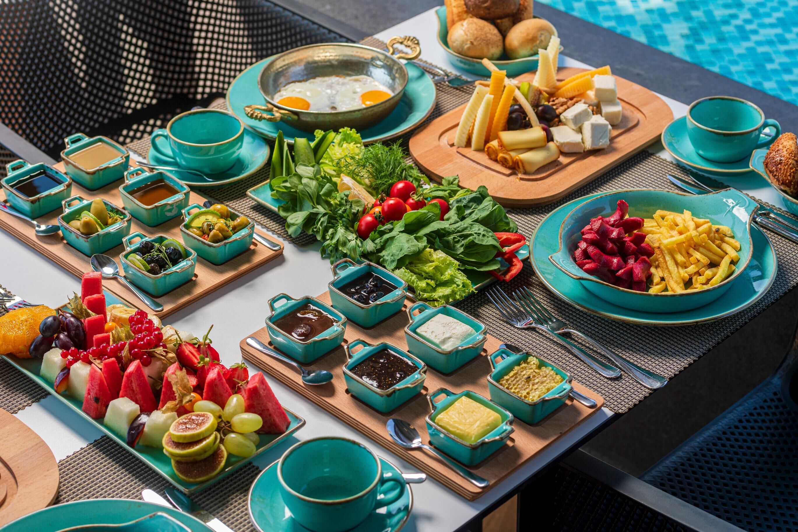 Una mesa con diferentes alimentos y condimentos, como frutas, verduras y queso, ubicada cerca de una piscina.