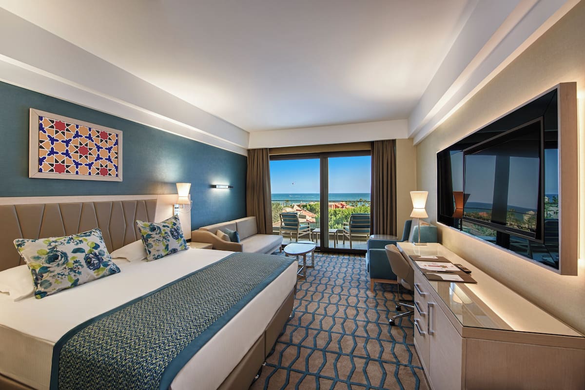 A hotel room with a bed, a desk, a TV, and a balcony with a view of the ocean.