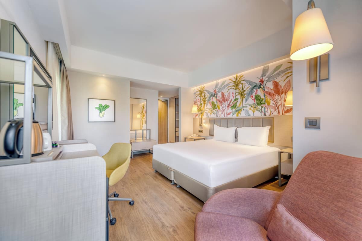 A modern hotel room with a large bed, a desk with a chair, a sofa, and a wall with a tropical mural.