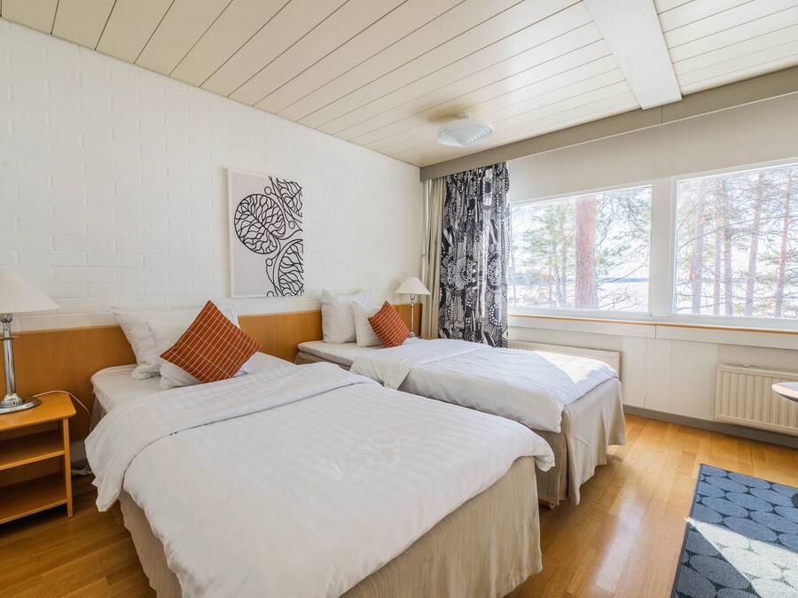Ein Hotelzimmer mit zwei Betten, einem Nachttisch, einer Lampe, einem Fenster mit Vorhängen und einem Heizkörper.