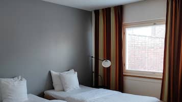 A hotel room with two single beds, a window with curtains, a ceiling light, and a view of buildings outside.