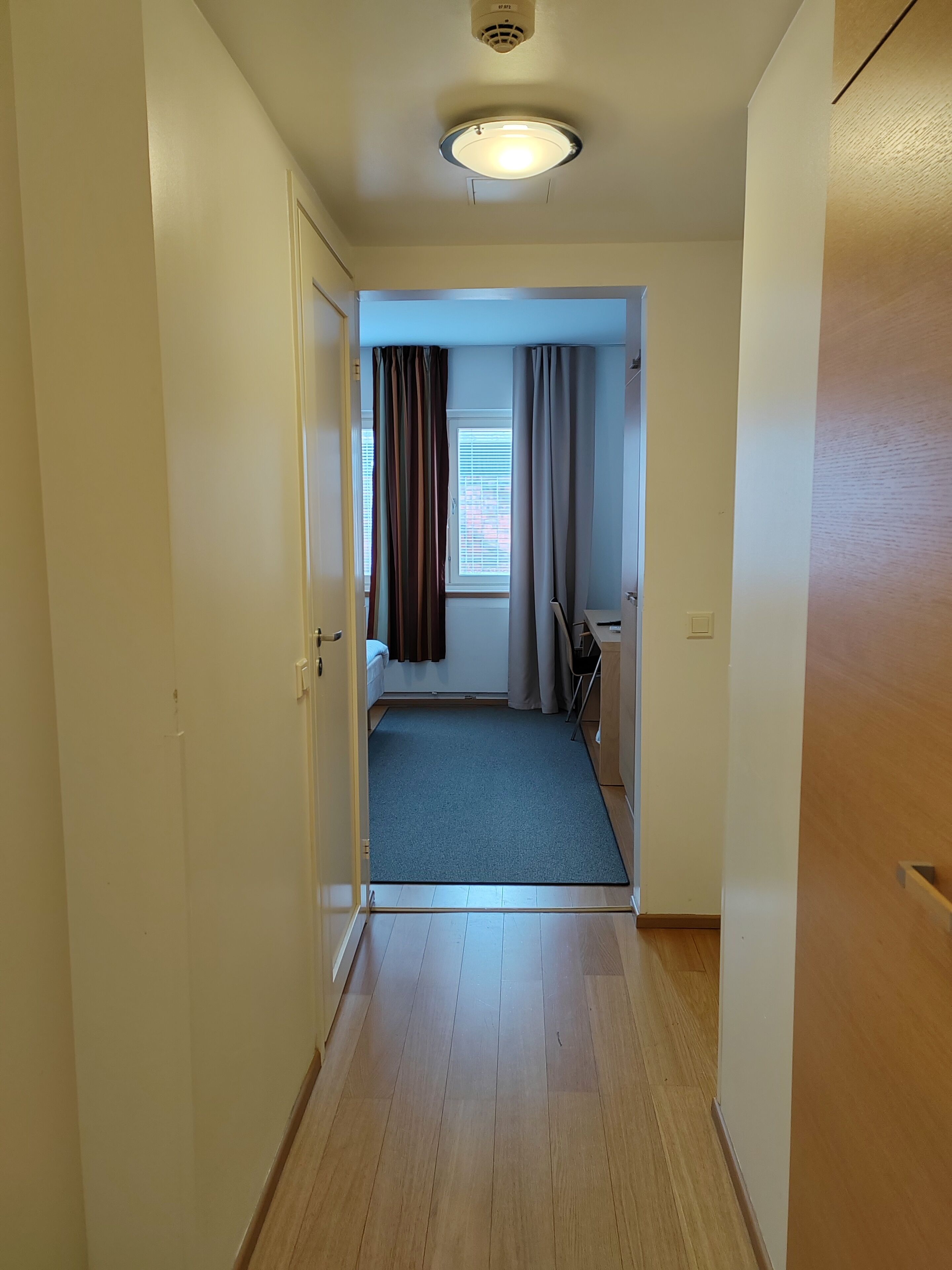 A narrow hallway with wooden flooring and white walls, leading to a room with a window, curtains, and a small table.