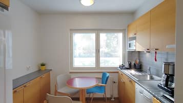 A compact kitchen with wooden cabinets, a dining table with chairs, a window with blinds, and a built-in dishwasher.
