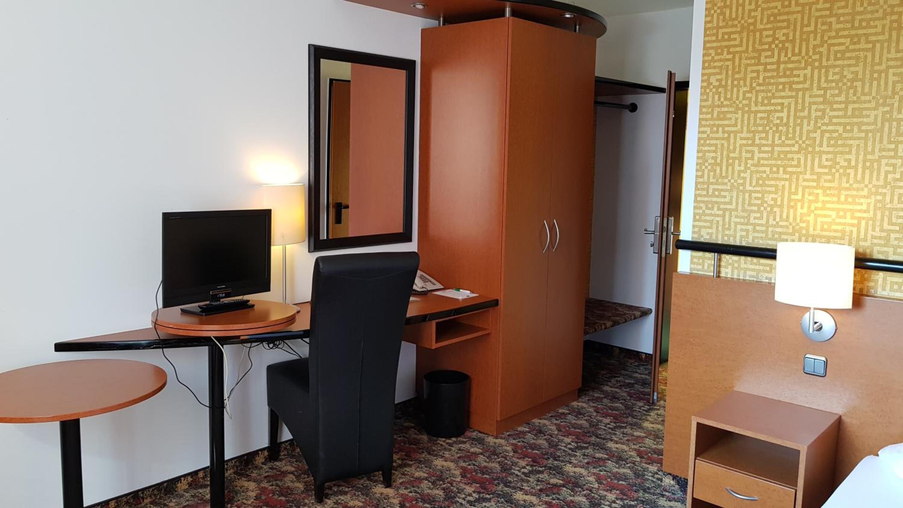 Et lille hotelværelse med skrivebord, fjernsyn, stol, garderobeskab og et natbord med lampe.