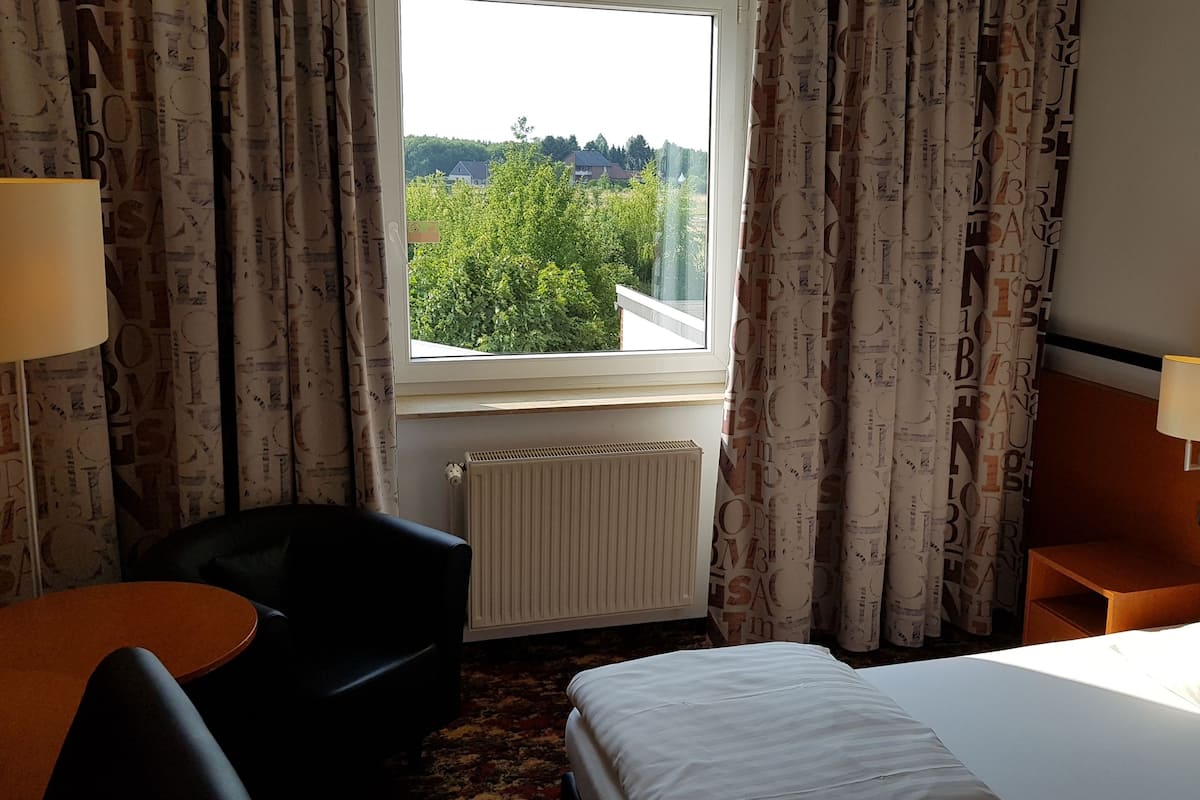 A hotel room with a bed, a chair, a small table, a lamp, and a view of greenery outside.