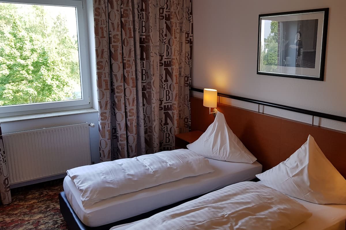 A hotel room with two single beds, a window with curtains, a wall-mounted picture, and two bedside lamps.