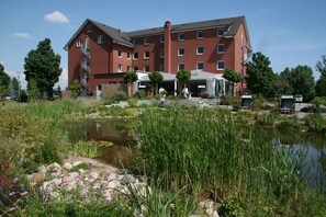 A multi-story brick building with a covered outdoor seating area and a pond. - Hotel & Restaurant Fricke (Lehrte)
