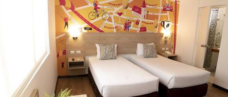 A hotel room with two single beds, a wooden desk, a potted plant, a ceiling fan, and a wall with a colorful map design.