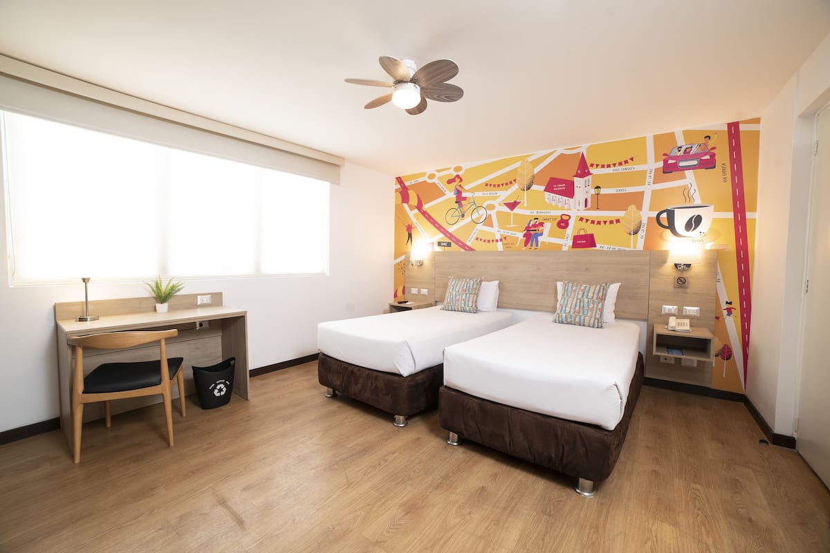 Ein Hotelzimmer mit zwei Betten, einem Schreibtisch, einem Stuhl, einem Deckenventilator, einem Fenster und einer Wand mit Weltkarten-Motiv.