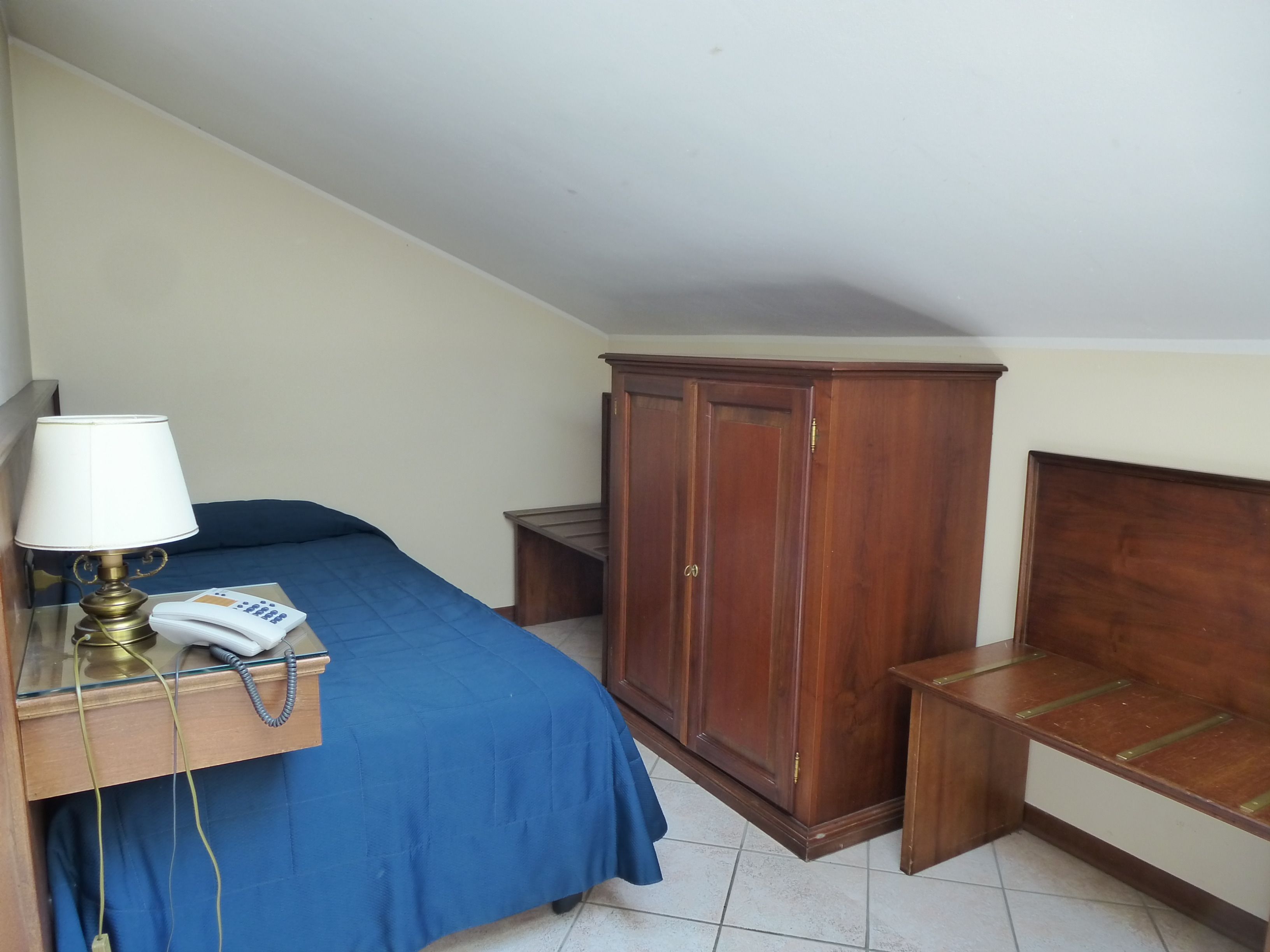 A bedroom with a bed, a wooden wardrobe, a bedside table with a lamp, and a telephone.
