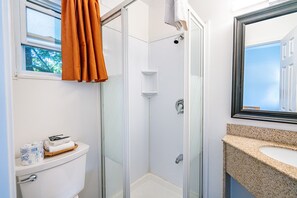 A bathroom with a glass shower enclosure, a toilet, a sink with a granite countertop, and a window with blinds.