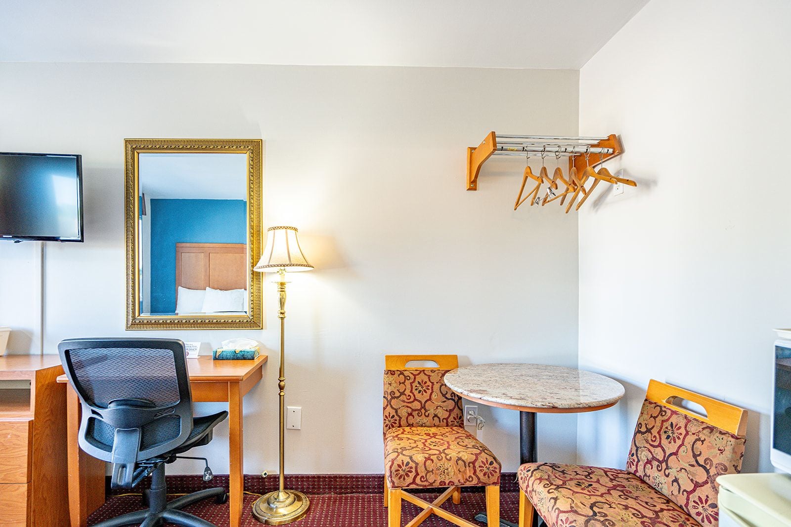 Une chambre d’hôtel équipée d’un bureau, d’une chaise, d’une table ronde, de chaises à motifs, d’une lampe, d’un miroir, d’une télévision et d’un porte-manteau.