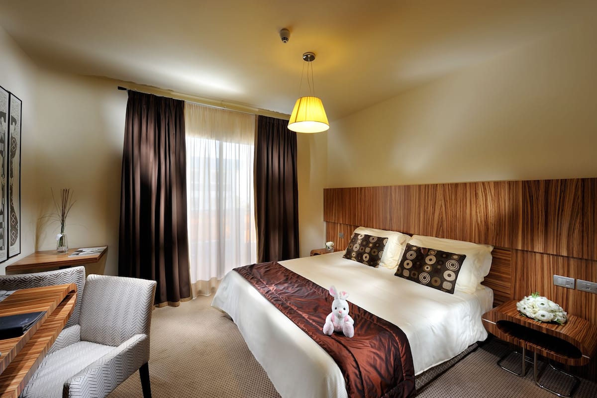 A hotel room with a large bed, a wooden headboard, a desk with a chair, and a window with curtains.