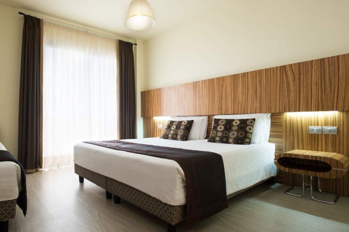 A hotel room with a large bed, a smaller bed, a wooden headboard, a round wooden table, and a large window with curtains.
