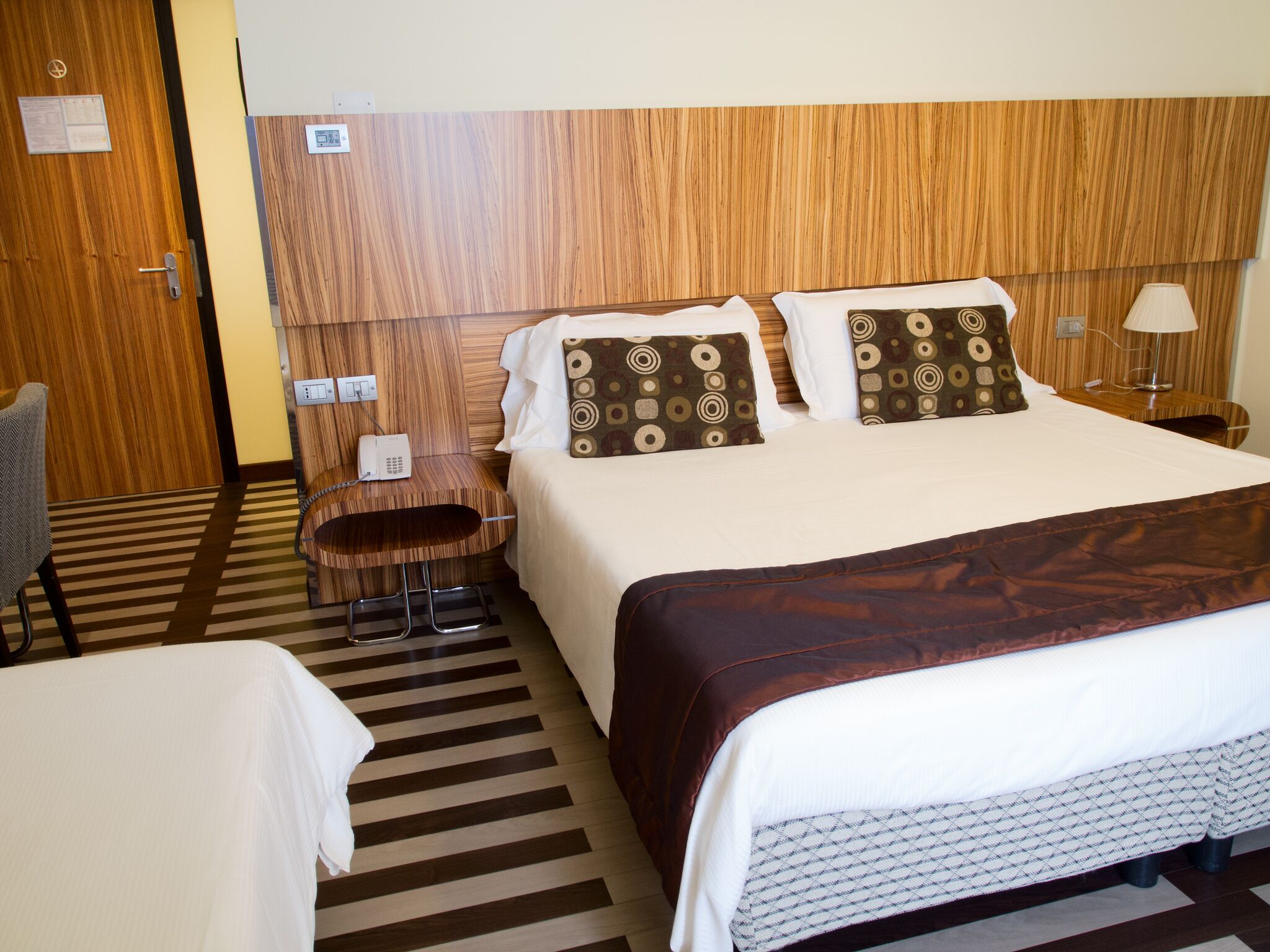 Een hotelkamer met een groot bed, twee kussens met ronde patronen, een nachtkastje met een lamp, een telefoon en een gestreept tapijt.