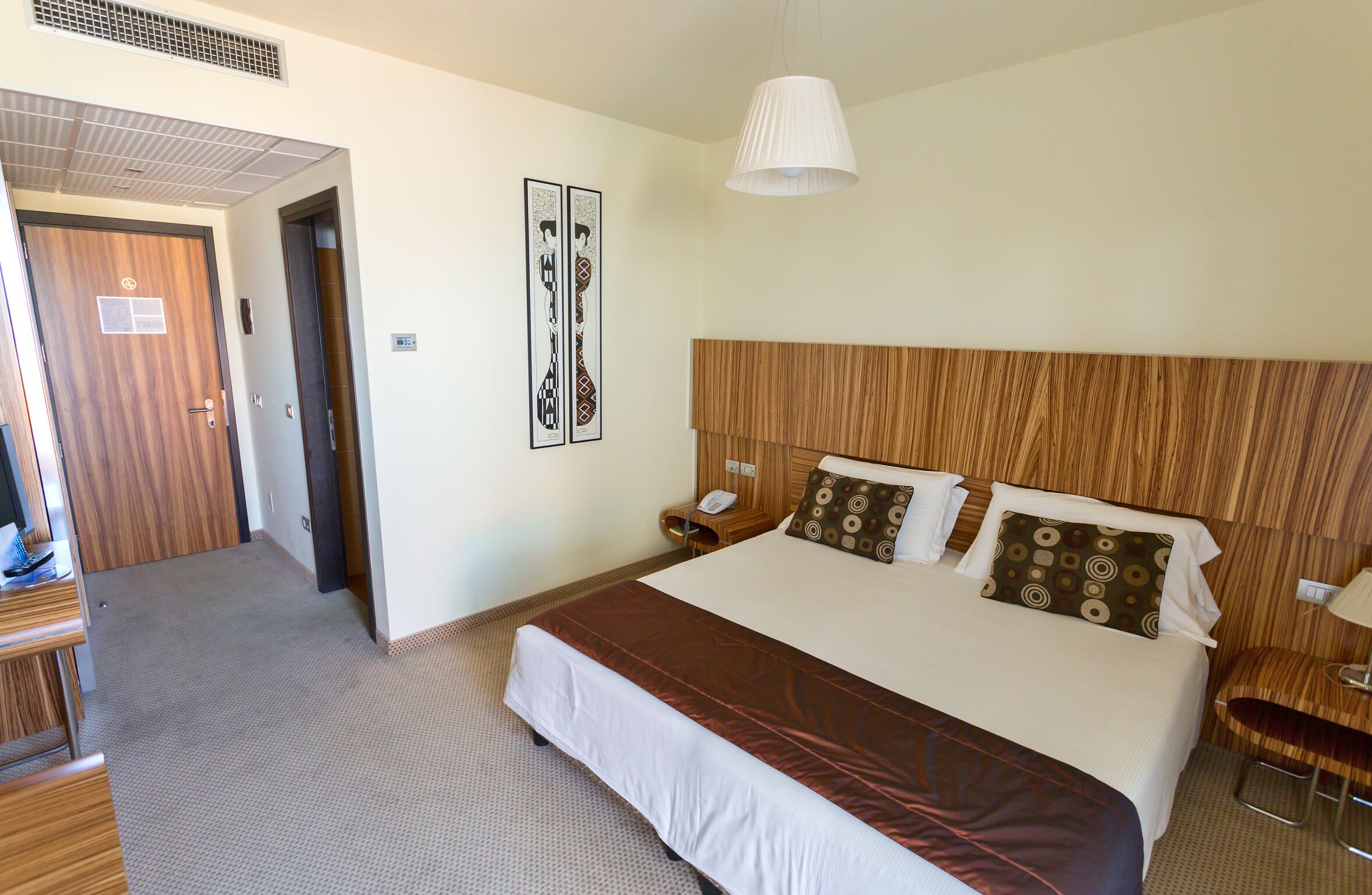 Habitación de hotel con una cama grande, cabecera de madera, una mesita de noche con lámpara y un teléfono en la pared.