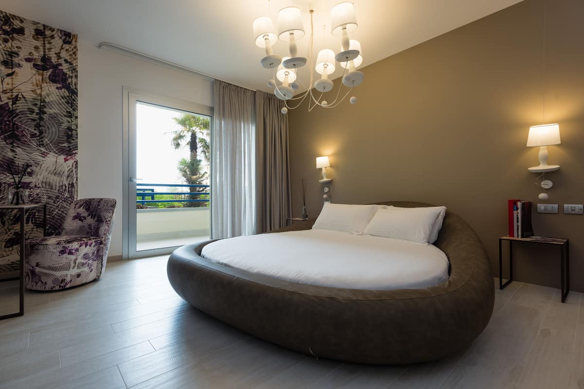 A modern bedroom with a large bed, a chandelier, a nightstand, and a view of the outdoors.