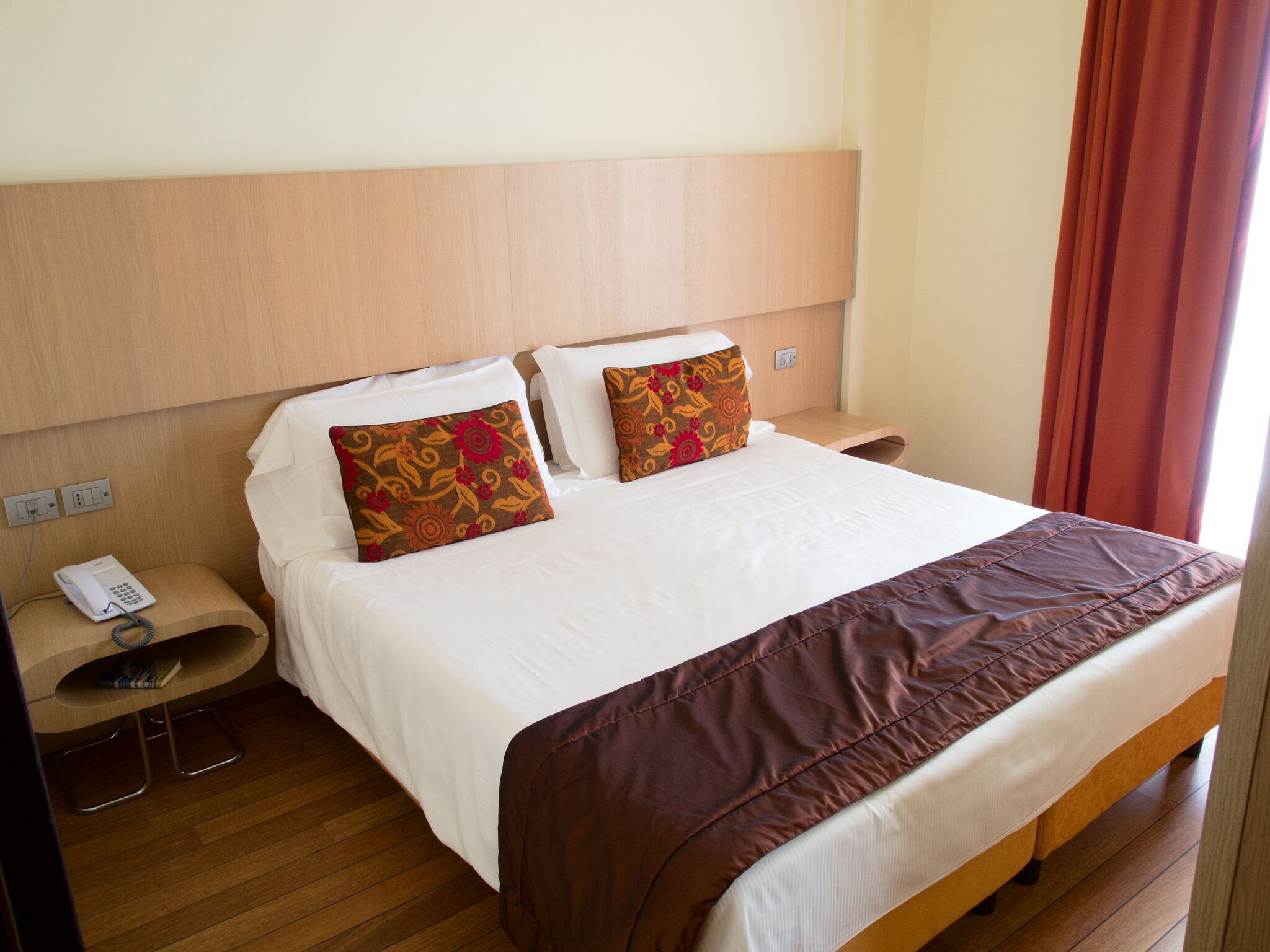 Een hotelkamer met een houten hoofdbord, een bed met witte lakens, twee decoratieve kussens, een nachtkastje met een telefoon en een rood gordijn.