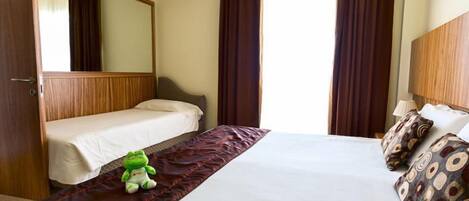 Habitación de hotel con una cama grande, una cama más pequeña, un espejo y dos ventanas con cortinas.