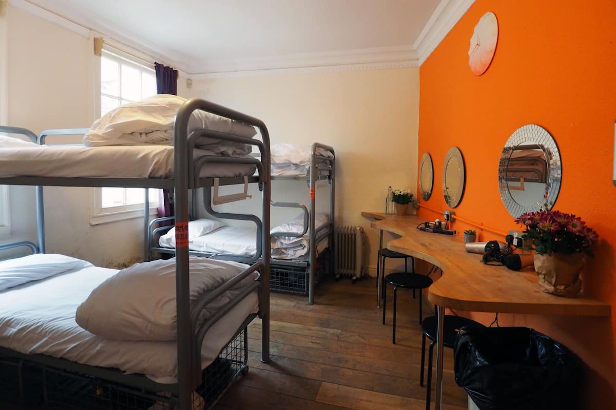 A dormitory room with bunk beds, a desk, a chair, a radiator, and a window.