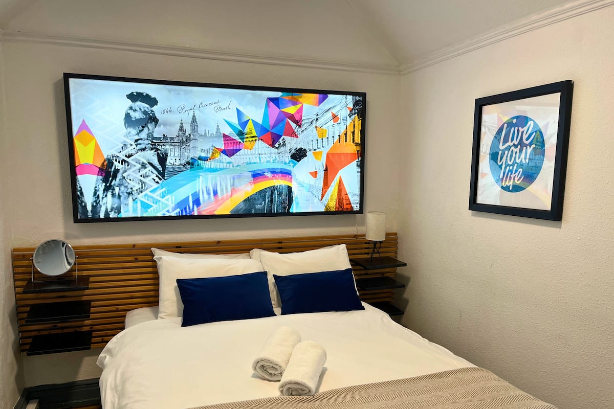 A hotel room with a bed, a large framed picture of a cityscape, a framed motivational quote, and a bedside table with a lamp.