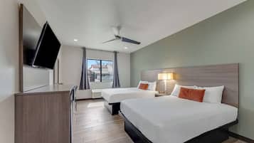 Habitación de hotel con dos camas, un televisor de pantalla plana montado en la pared, ventilador de techo y una ventana con cortinas.