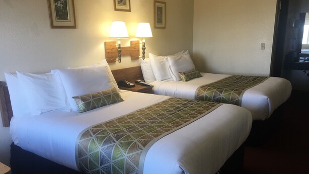 A hotel room with two beds, each with white linens and geometric-patterned bedspreads. There are two bedside lamps and a nightstand with a phone and a lamp. - Westward Inn (Brookings)