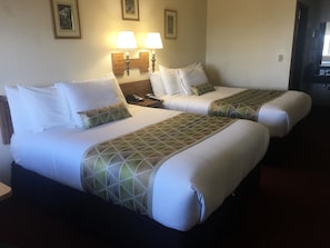 Quarto de hotel com duas camas, cada uma com roupa de cama branca e colchas com padrão geométrico. Há duas luminárias de cabeceira e uma mesa de cabeceira com um telefone e uma luminária.