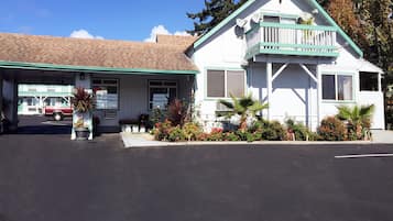 A white building with a green roof and a balcony, surrounded by potted plants and flowers.
