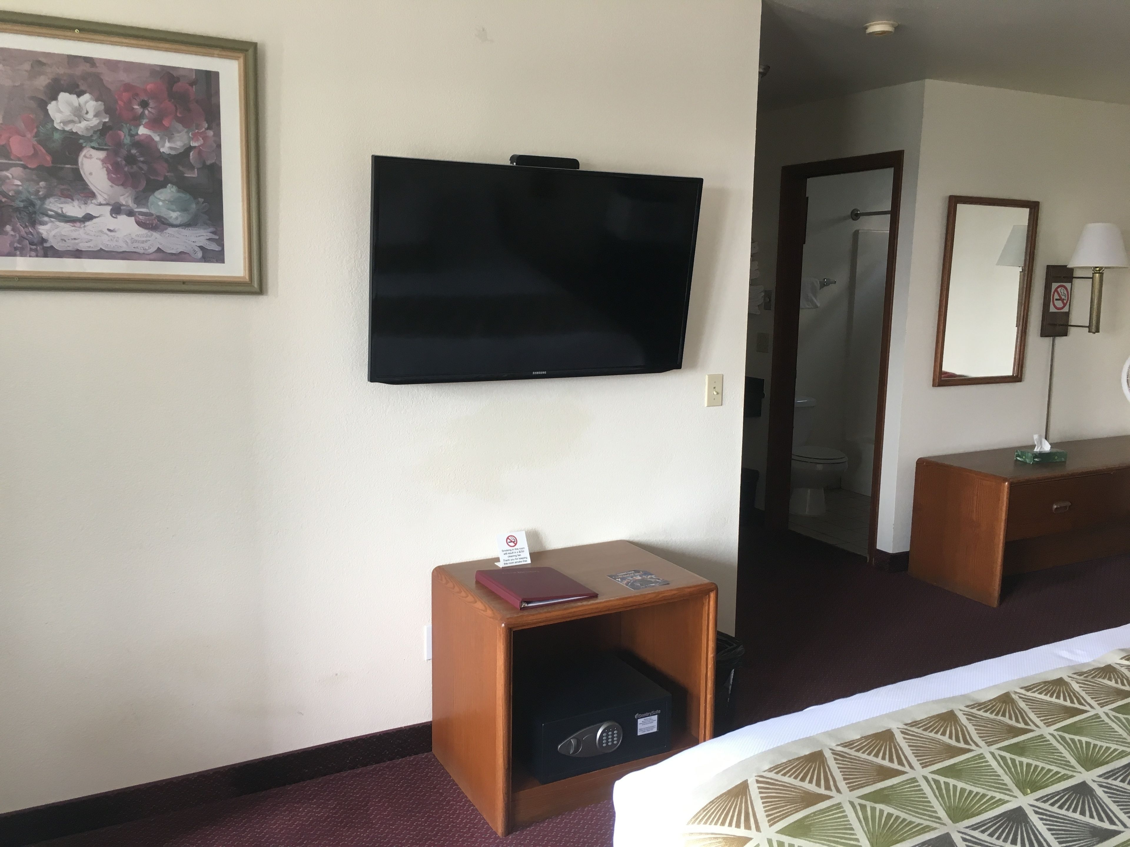 A hotel room with a flat-screen TV mounted on the wall, a small wooden side table with a book and a remote control, a bed with a patterned bedspread, and a bathroom visible through an open door.