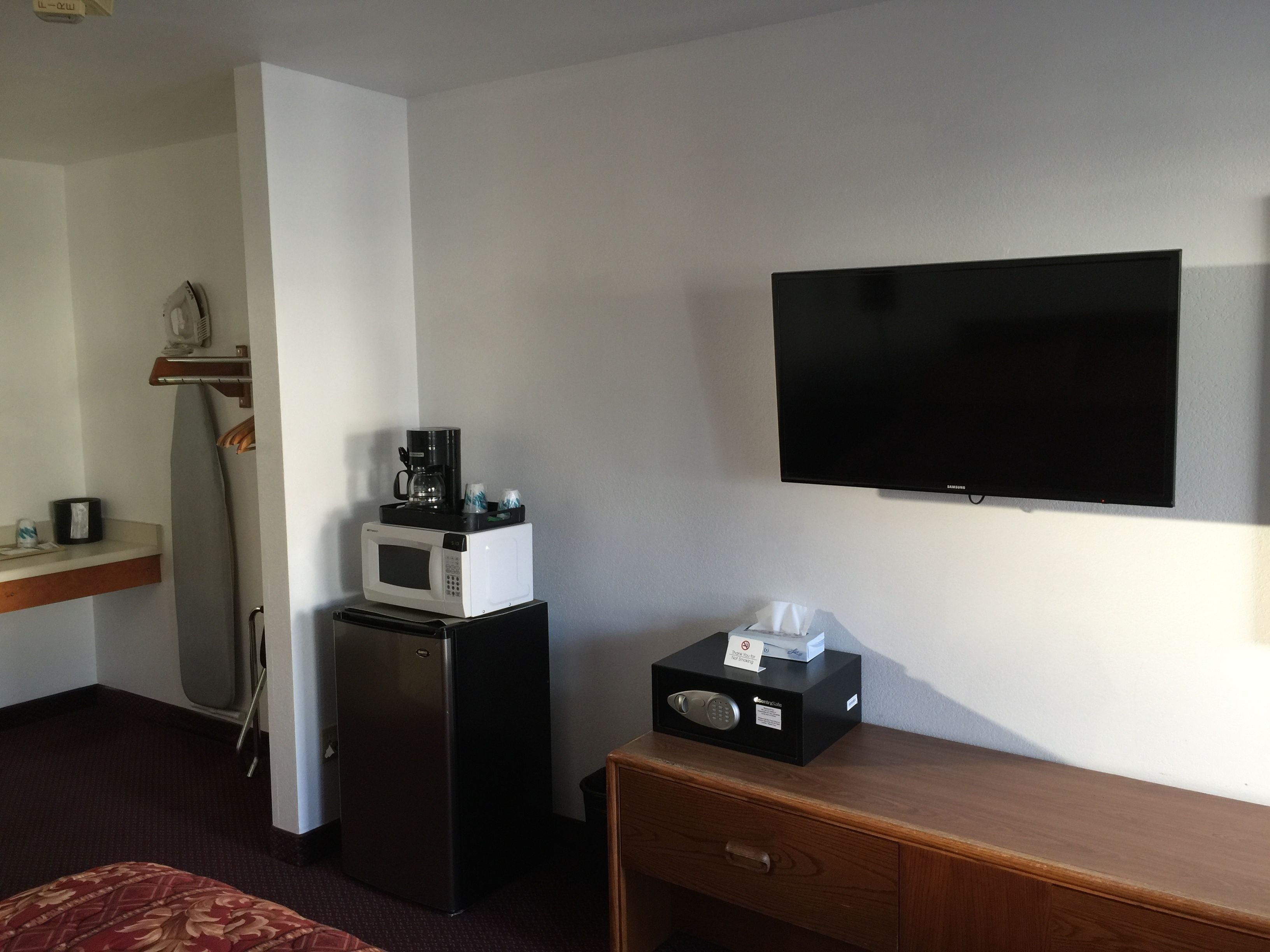 A hotel room with a flat-screen TV mounted on the wall, a microwave on a small table, a refrigerator, a wooden desk with a drawer, and a bed with a patterned bedspread.