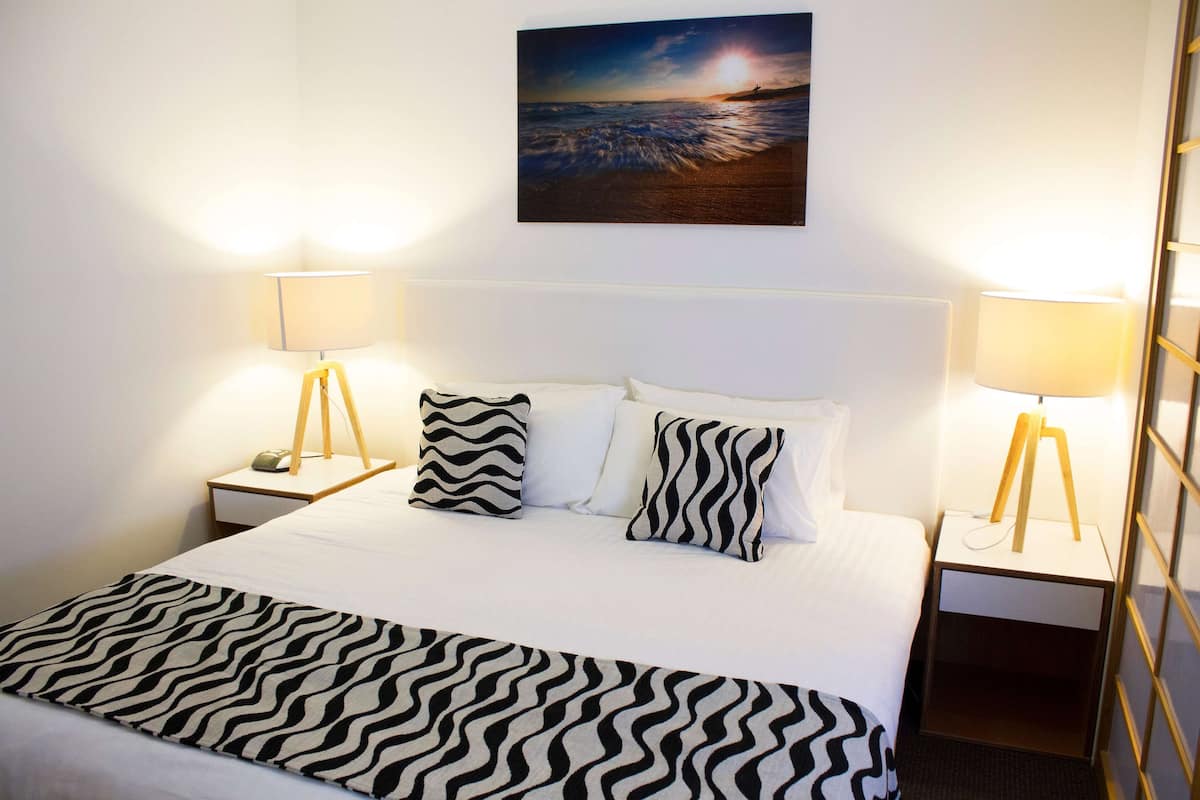Ein ordentlich bezogenes Bett mit einem zebra-gemusterten Überwurf, zwei Nachttischlampen und einem an der Wand hängenden Bild von einem Sonnenuntergang über dem Meer.