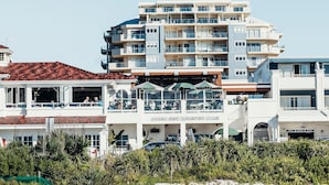 Een hotel aan het strand met meerdere balkons, een zandstrand en een helderblauwe lucht.