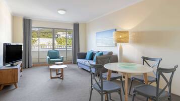 Habitación de hotel con un ventanal grande, un sofá, un televisor y un comedor con sillas y una mesa.