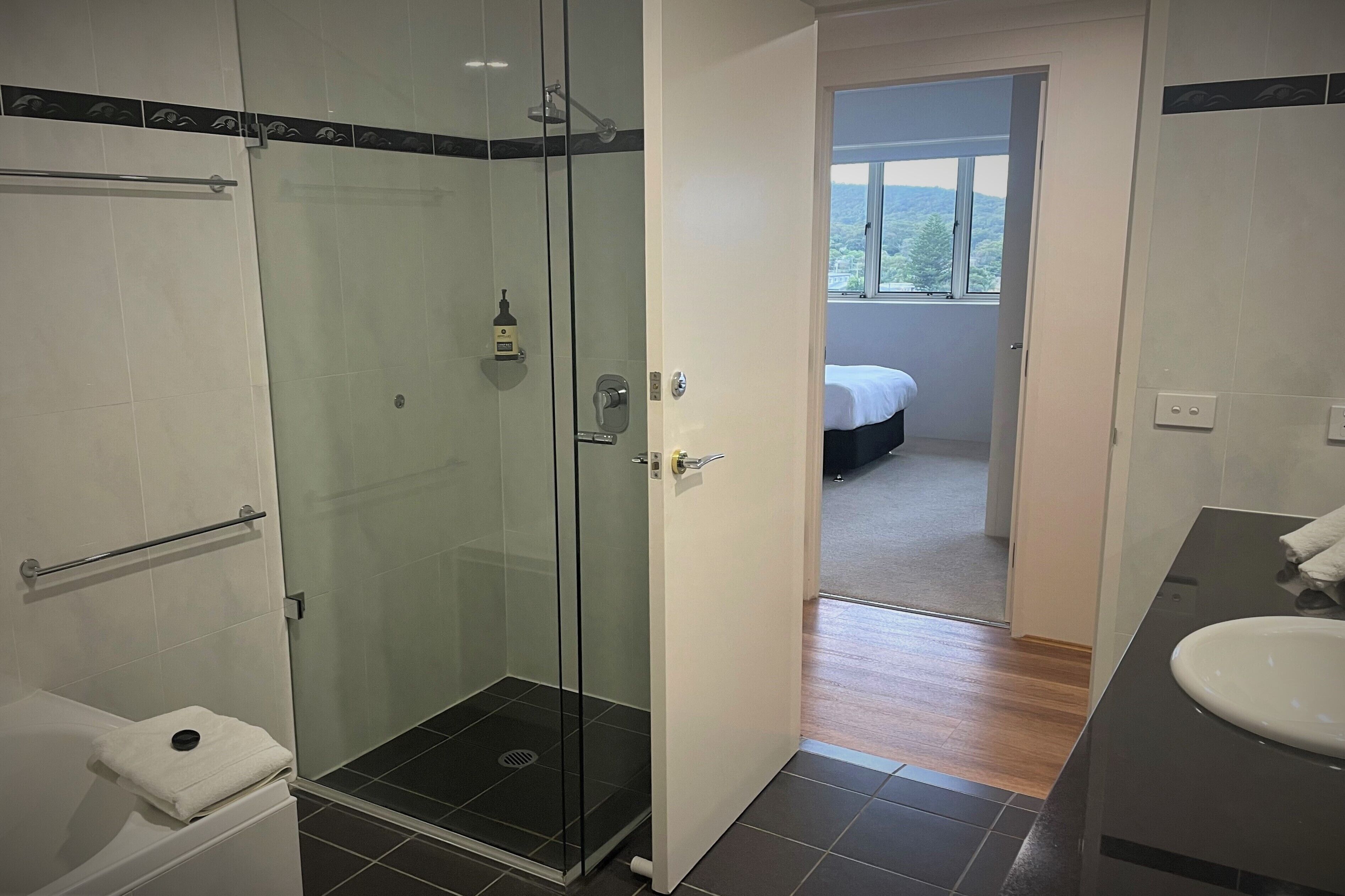 Une salle de bain moderne avec une douche à parois de verre, un lavabo blanc et une chambre à coucher visible à travers une porte ouverte.