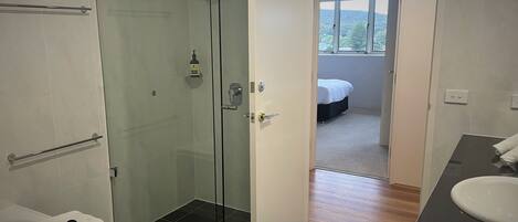 A modern bathroom with a glass-enclosed shower, a white sink, and a visible bedroom through an open door.