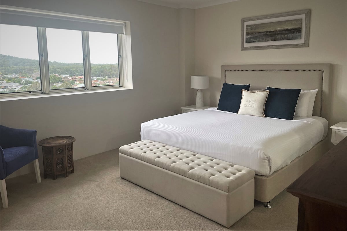 Ein Schlafzimmer mit einem großen Bett, einem Nachttisch mit Lampe, einem kleinen runden Beistelltisch, einer Bank, einem Sessel und einem Fenster mit Landschaftsblick.