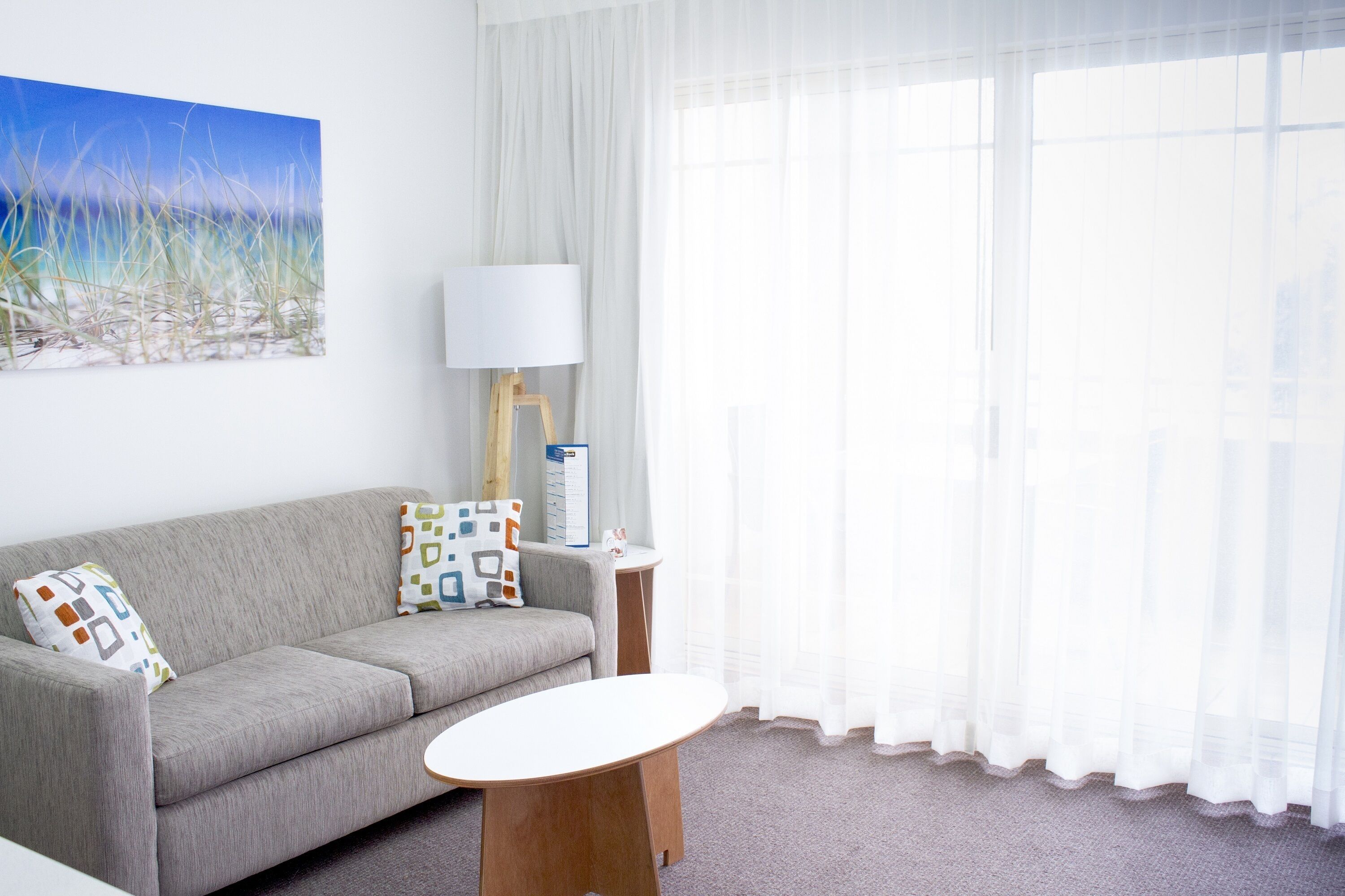 A modern living room with a grey sofa, a small round table, a lamp, and a painting of a beach scene.