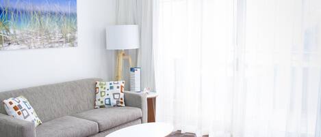 A modern living room with a grey sofa, a small round table, a lamp, and a painting of a beach scene.
