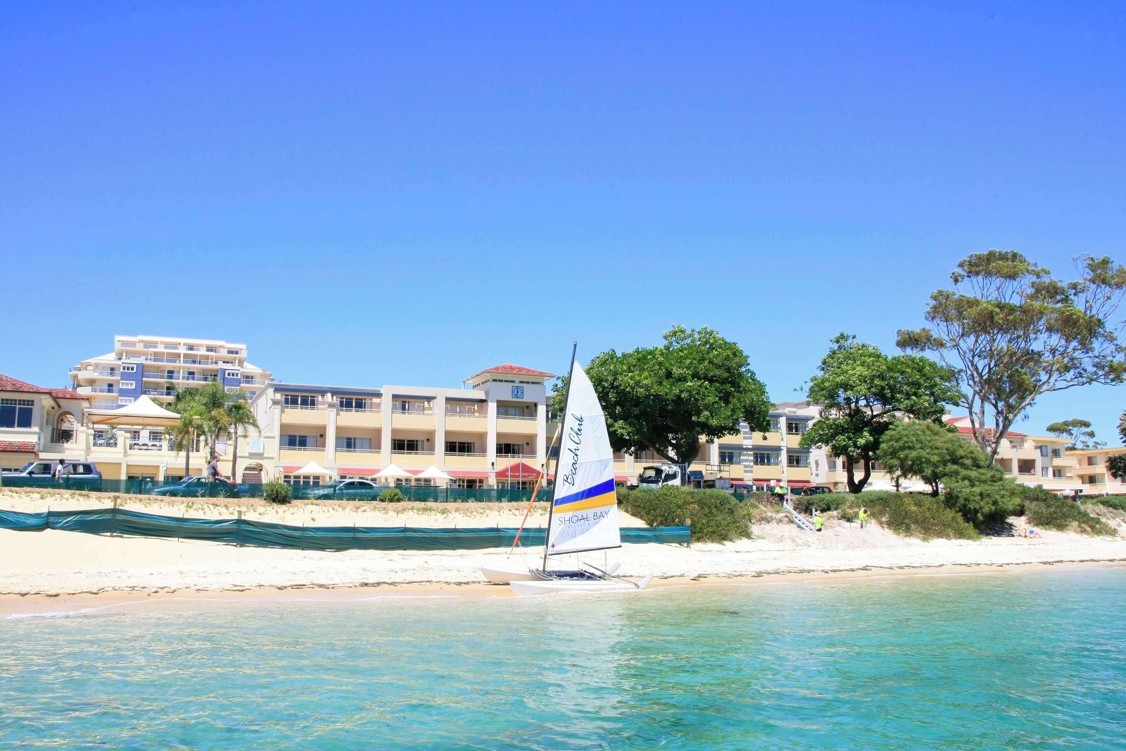 Um hotel à beira-mar com um veleiro na água, praia de areia e edifícios ao fundo.