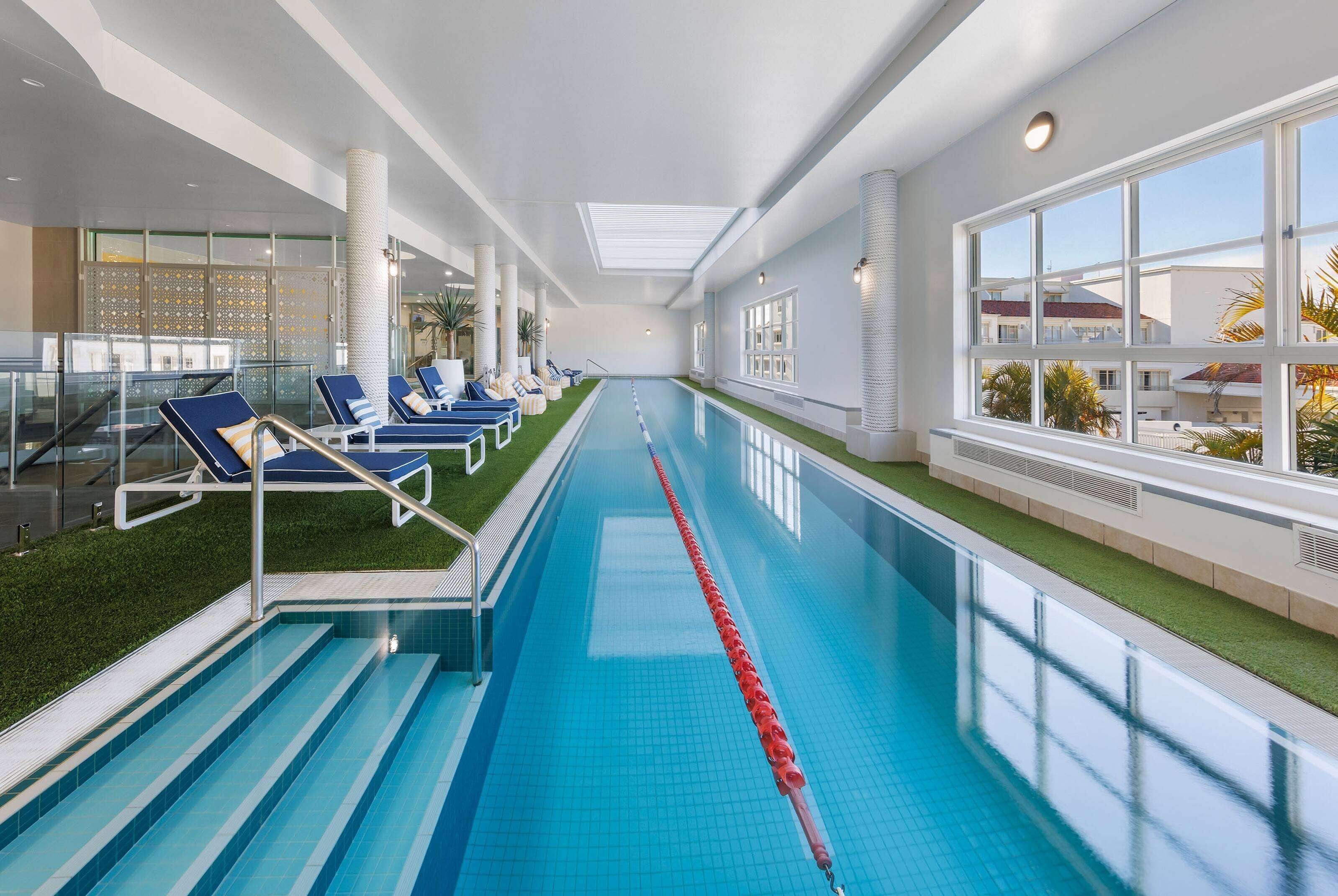 Una piscina interior con azulejos azules, un separador de carril rojo y sillones de descanso a los lados.